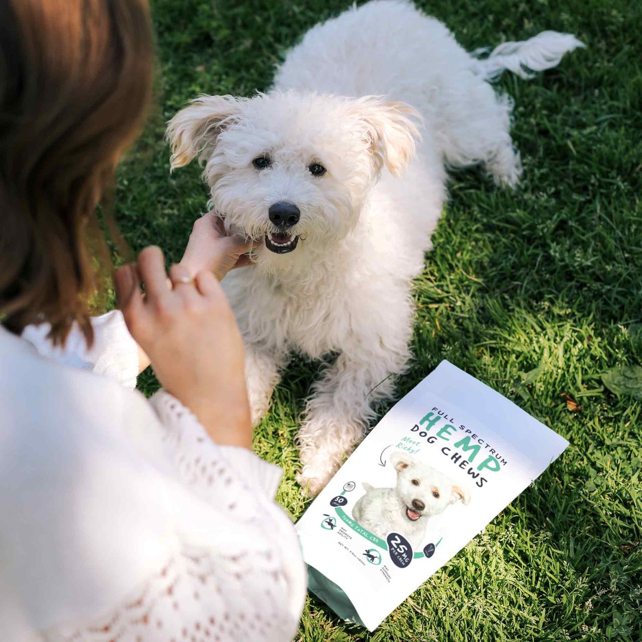 Fluffy white dog lying on grass with a person petting it. Hemp Dog Chews pouch placed beside the dog.