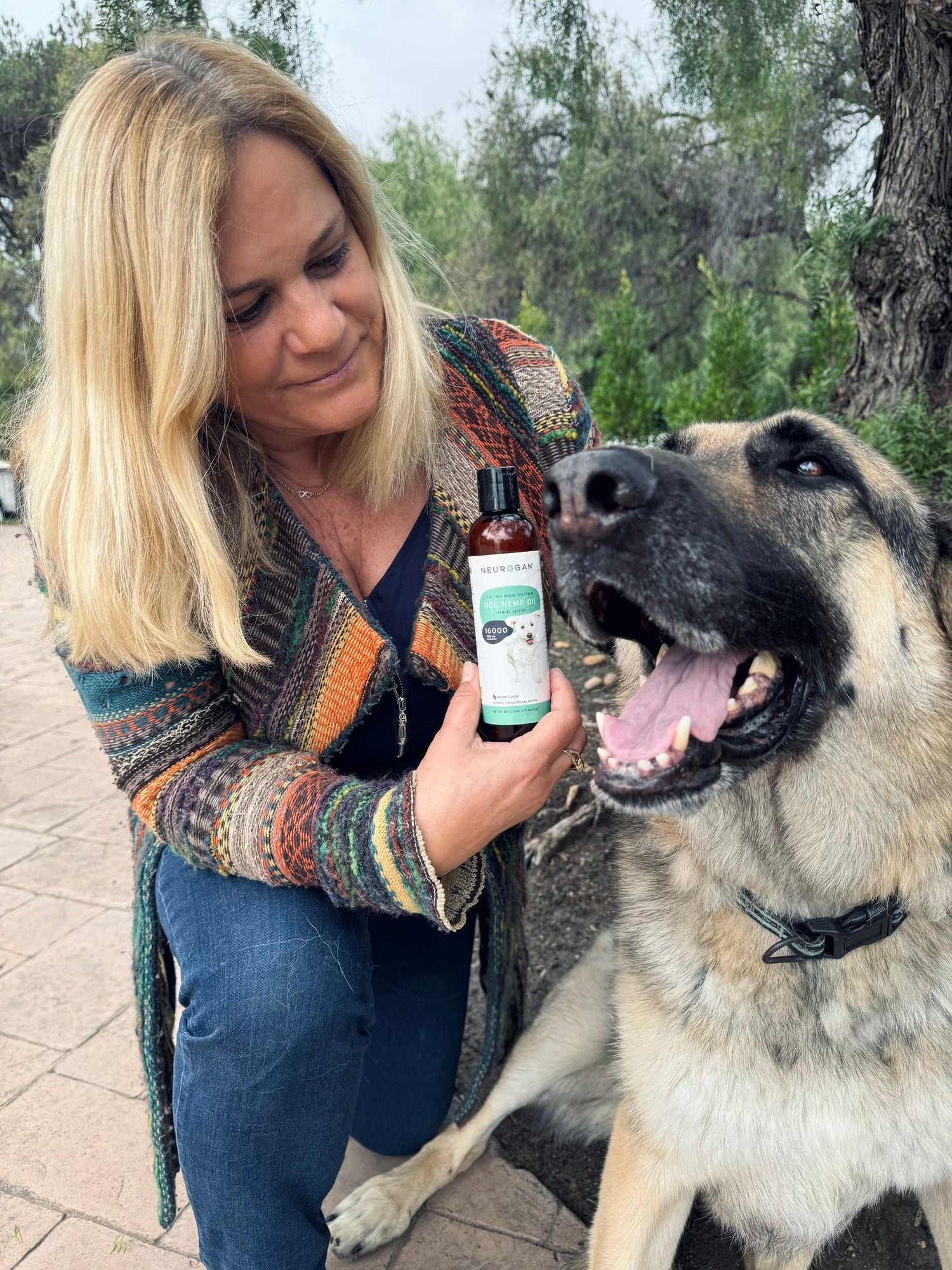 Smiling woman holding Dog Hemp Oil bottle next to a happy German Shepherd outdoors.
